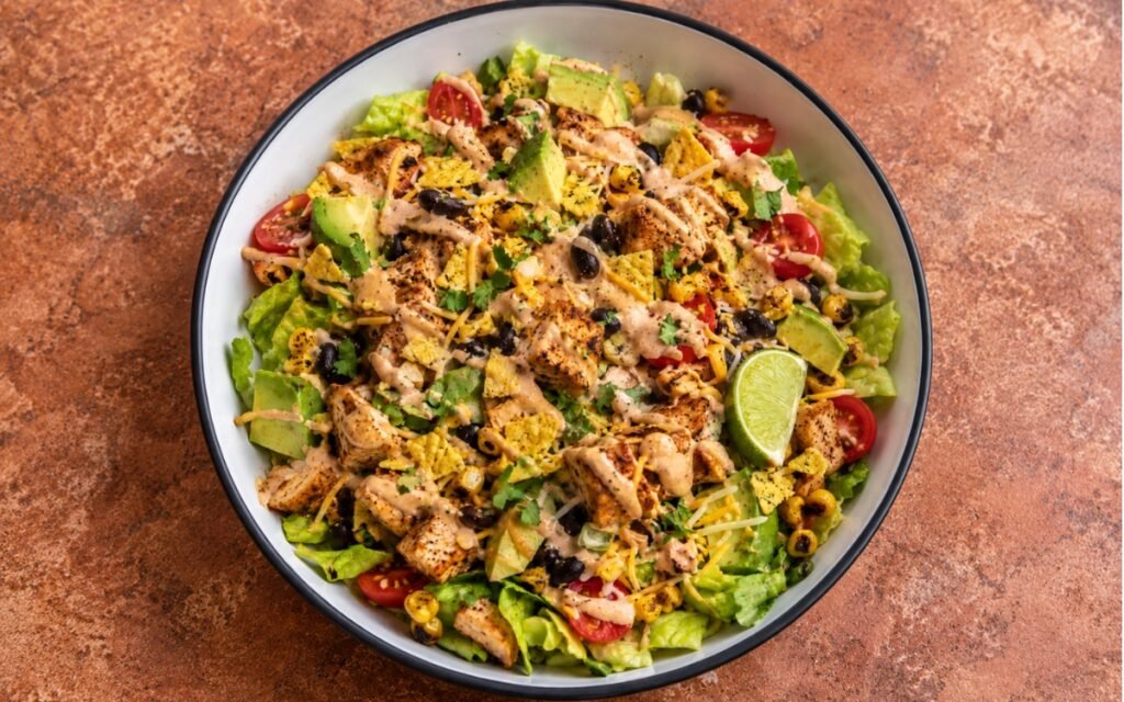 Mexican chicken taco salad showing seasoned chicken, black beans, charred corn, avocado, and crushed tortilla chips with chipotle crema, on marble surface