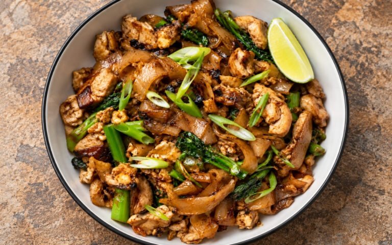 Pad See Ew showing glossy, lightly charred wide rice noodles with Chinese broccoli, chicken, and egg, on marble surface