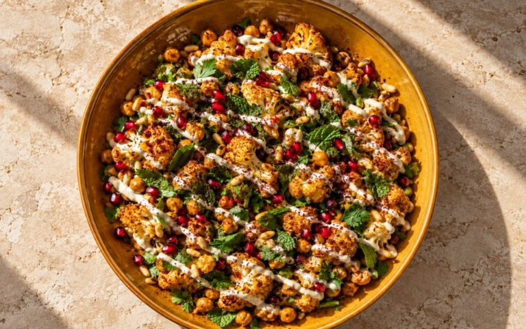 Roasted cauliflower and chickpea salad showing deeply golden roasted vegetables with thick tahini drizzle, pomegranate seeds, and fresh herbs, on marble surface