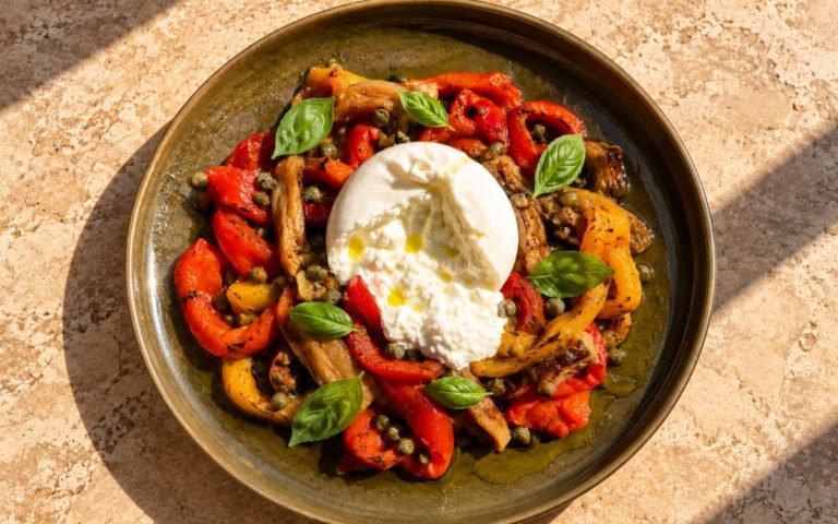 Spanish escalivada showing soft roasted pepper and eggplant strips with torn creamy burrata, capers, and fresh basil, on marble surface