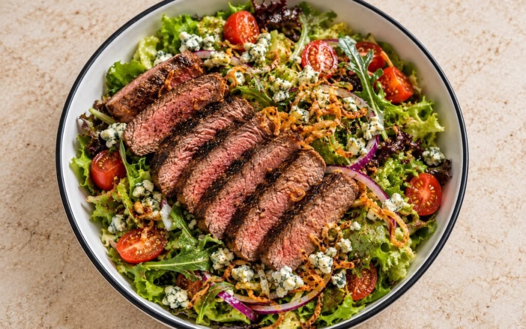 Steakhouse steak salad showing sliced medium-rare steak over crisp greens with blue cheese crumbles and crispy fried shallots, on marble surface