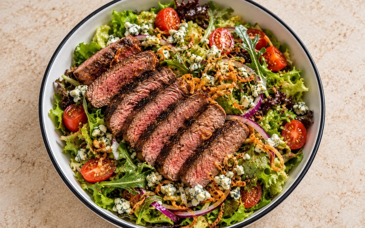 Steakhouse steak salad showing sliced medium-rare steak over crisp greens with blue cheese crumbles and crispy fried shallots, on marble surface