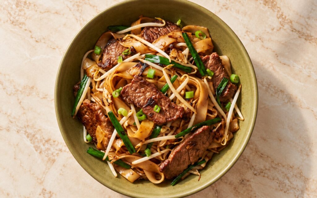 Beef chow fun showing glossy wide rice noodles with wok-charred patches, tender beef slices, bean sprouts, and Chinese chives in dark soy sauce, on marble surface