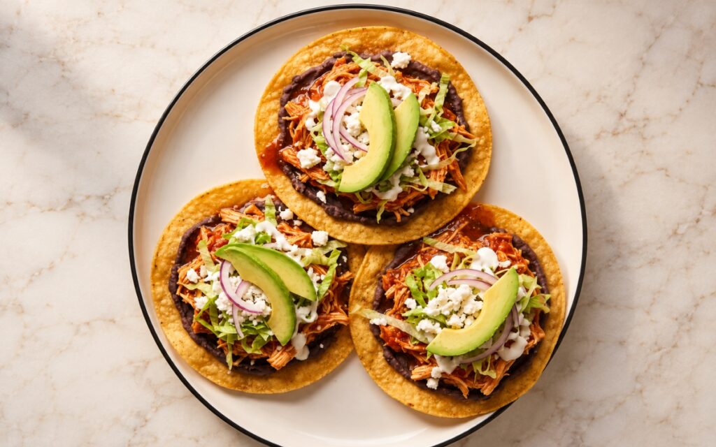 Chicken tinga tostadas showing crisp golden corn shells topped with black beans, glossy red chipotle chicken, lettuce, queso fresco, crema, and avocado, on marble surface