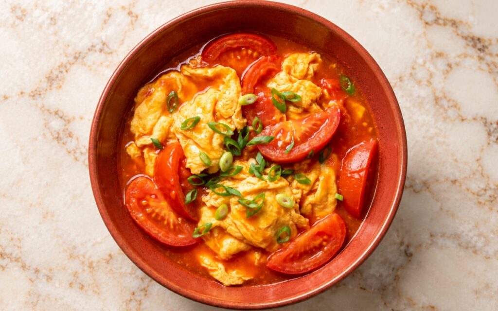 Chinese tomato egg stir-fry showing large soft scrambled egg curds, juicy red tomato wedges in a glossy sauce, and sliced scallion greens, on marble surface