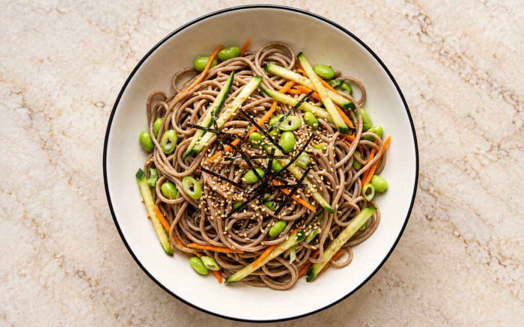 Cold soba noodle salad showing loose tangles of buckwheat noodles with cucumber and carrot matchsticks, edamame, scallions, sesame seeds, and fine nori strips in a white bowl, on marble surface