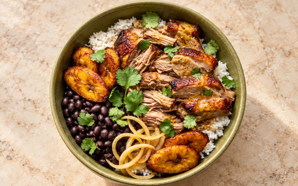 Cuban mojo pork rice bowl showing shredded and sliced citrus-garlic pork over white rice with black beans, sautéed onions, fried sweet plantains, and cilantro, on marble surface