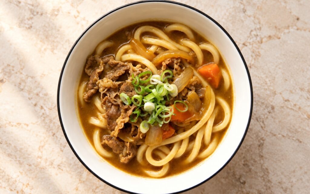 Japanese curry udon showing thick chewy udon noodles in glossy golden-brown curry broth with thin-sliced beef, soft onion, carrot, and sliced scallion, on marble surface