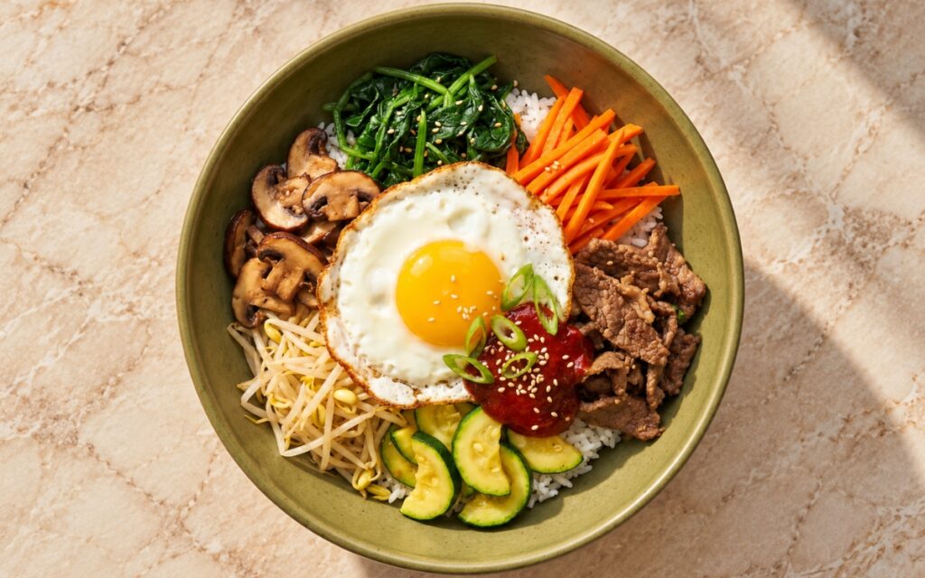 Korean bibimbap showing white rice topped with separate sections of spinach, carrots, mushrooms, bean sprouts, zucchini, and browned beef, a sunny-side-up egg, and gochujang, on marble surface