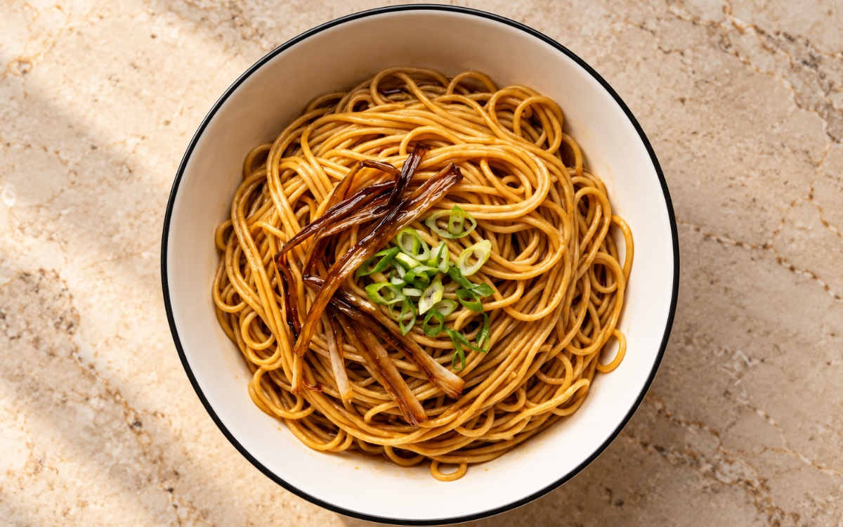 Shanghai scallion oil noodles showing a glossy amber mound of thin wheat noodles topped with dark golden fried scallion strands and sliced green onion, on marble surface
