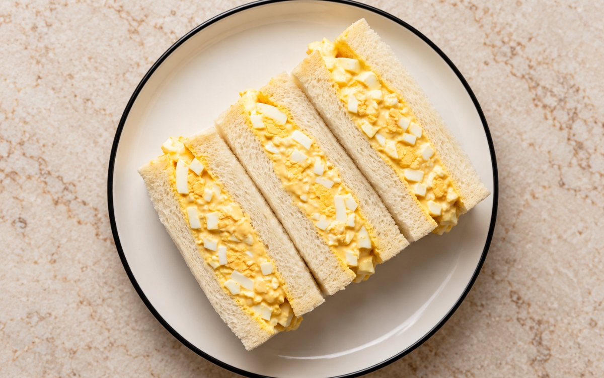 Tamago sando showing crustless milk bread halves stacked upright with thick creamy egg filling and visible chopped egg whites, on marble surface