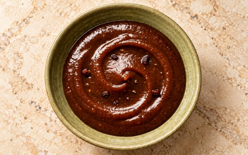 Tamarind date chutney showing a glossy dark brown sauce in a small bowl, a spoon trailing a thick ribbon, and thin date slices on top, on marble surface