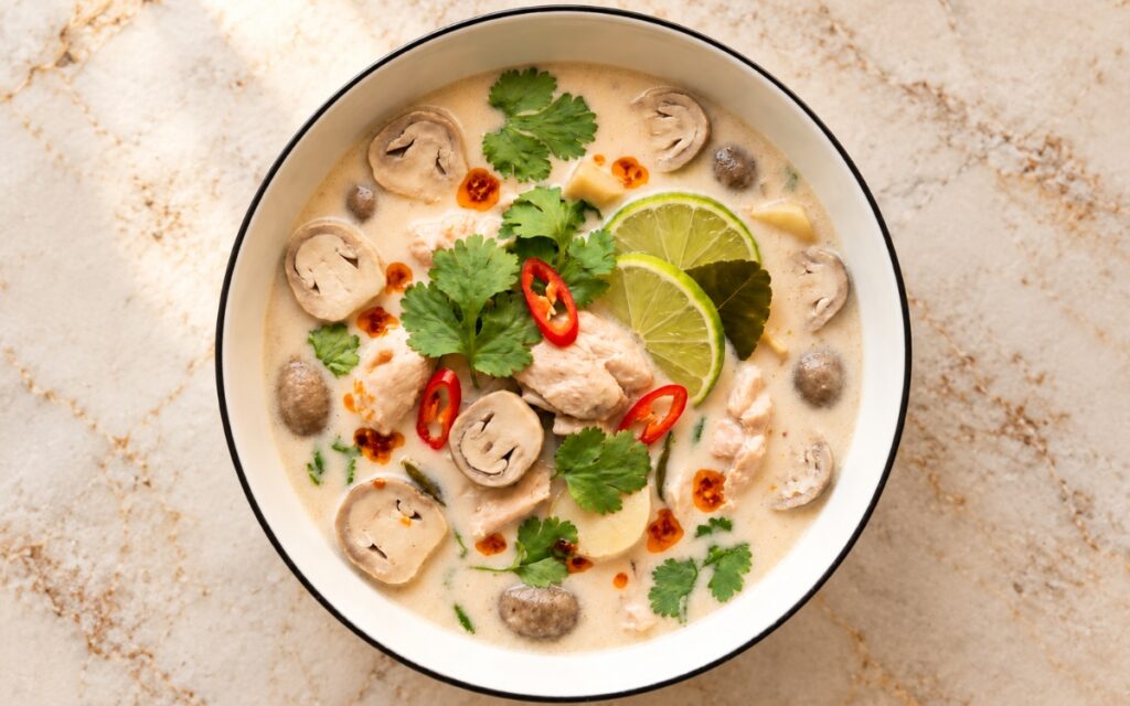 Tom Kha Gai Coconut Chicken Soup showing chicken and mushrooms in a pale ivory coconut broth with cilantro, sliced red chilli and lime wedges, on marble surface